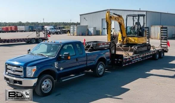 A blue flatbed tow truck carrying a yellow mini excavator at an industrial facility with shipping containers in the background.