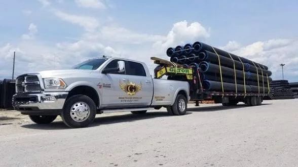 A white commercial pickup truck towing a large flatbed trailer loaded with industrial pipes under a cloudy sky.