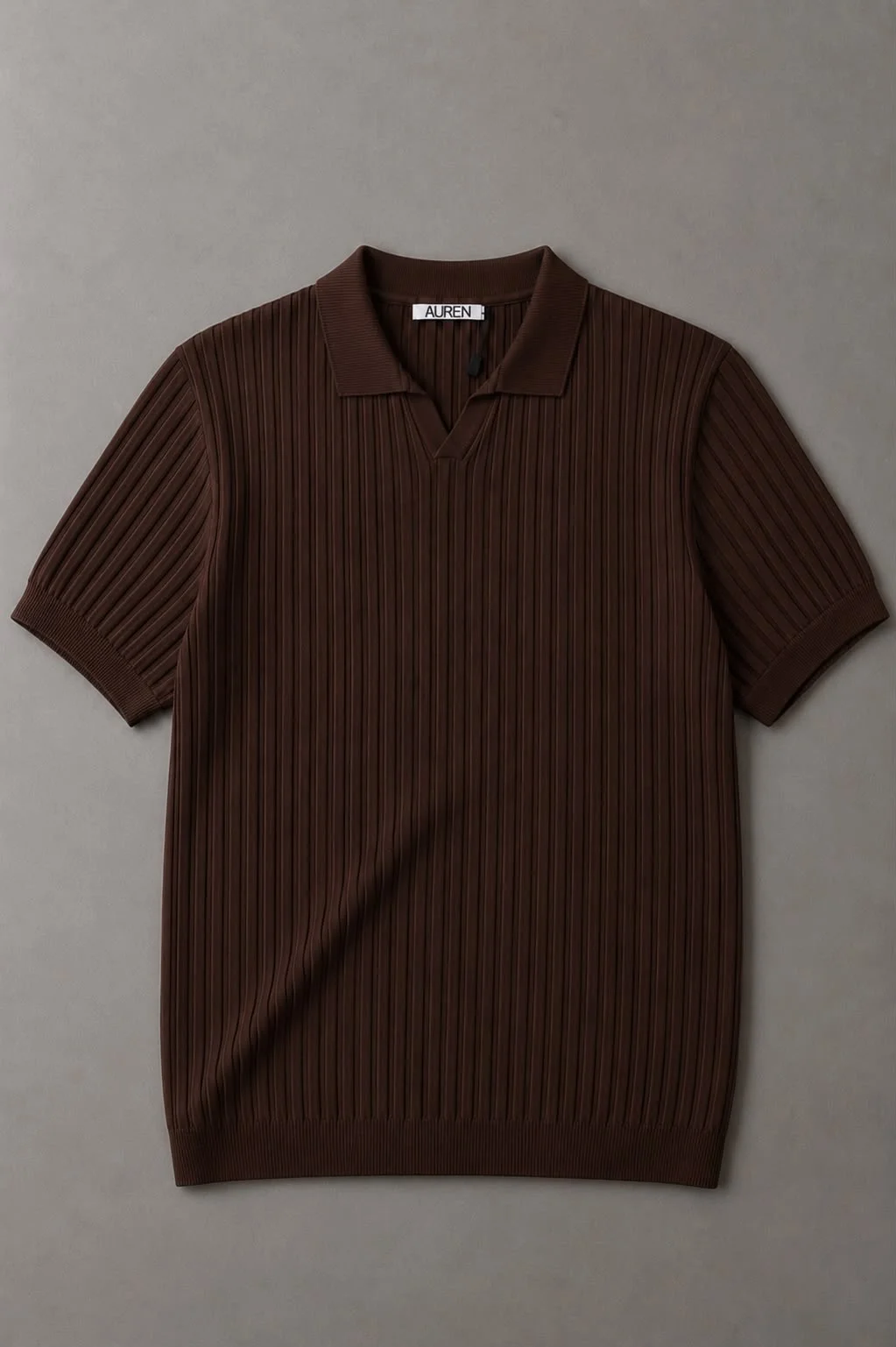 Polo de malha marrom escuro com textura plissada vertical, mangas curtas, gola clássica e etiqueta AUREN, fotografado sobre fundo cinza neutro.
