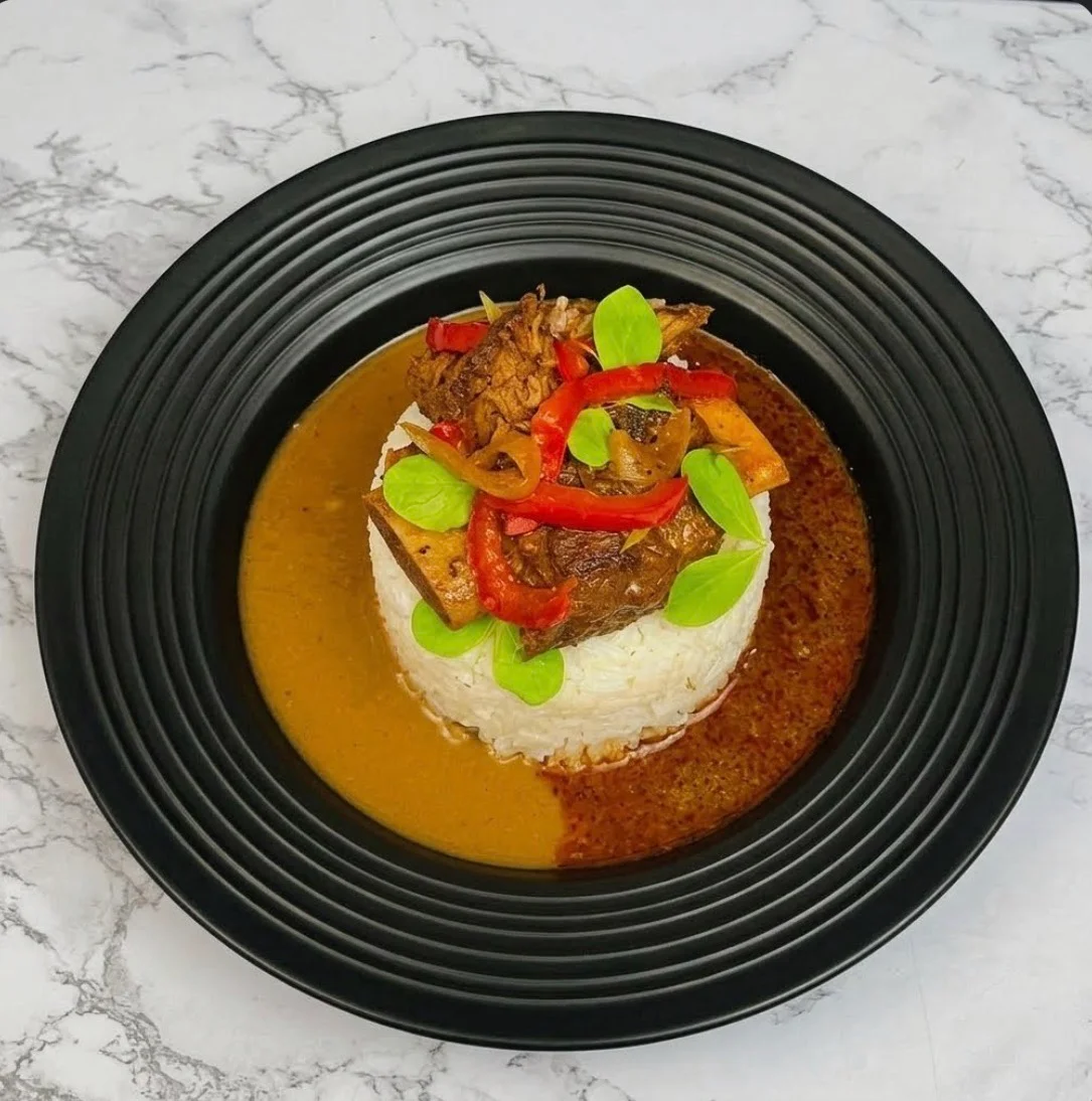Rice topped with saucy pieces, red pepper strips and fresh green leaves, with two sauces on a black plate.
