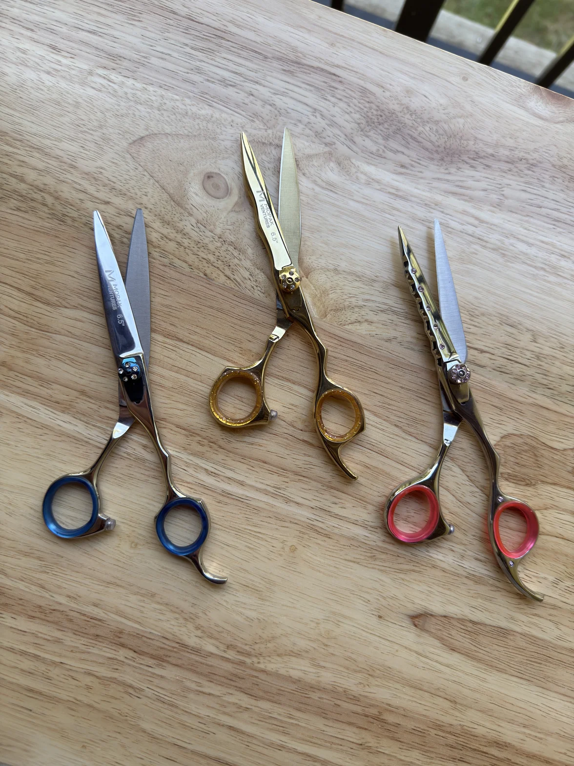 Three professional hairdressing scissors arranged on a light wooden table, including blue, gold, and red-handled shears with polished metal blades