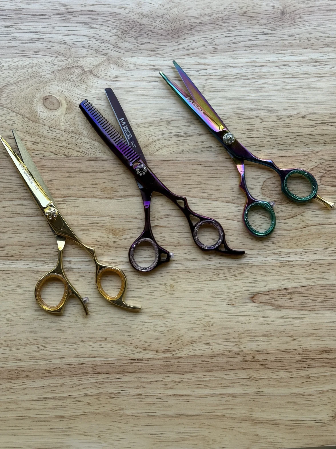Three colorful professional hairdressing scissors, including a thinning shear, arranged on a light wood surface