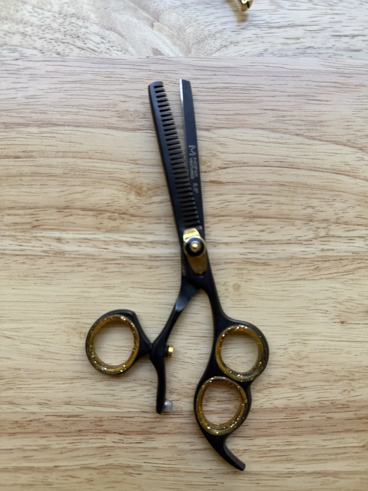 Black professional hairdressing scissors with thinning comb teeth and gold finger rings on a light wood surface