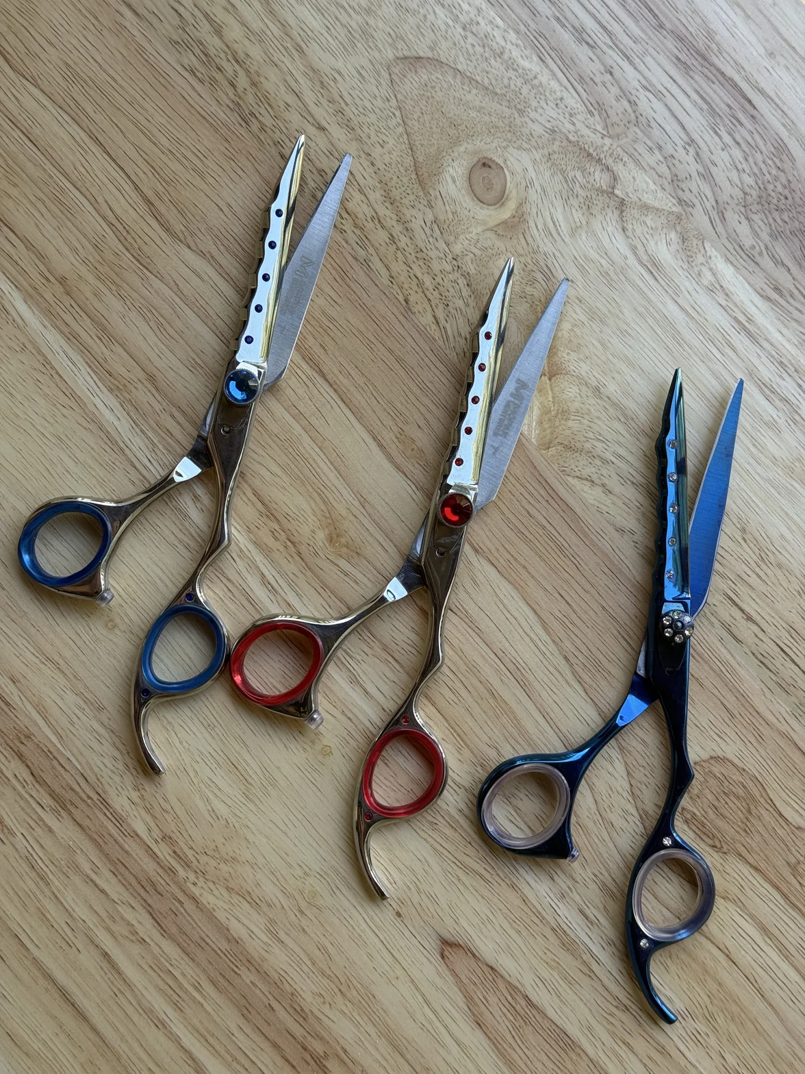 Several professional hairdressing scissors in silver, red, and blue finishes arranged on a light wooden surface