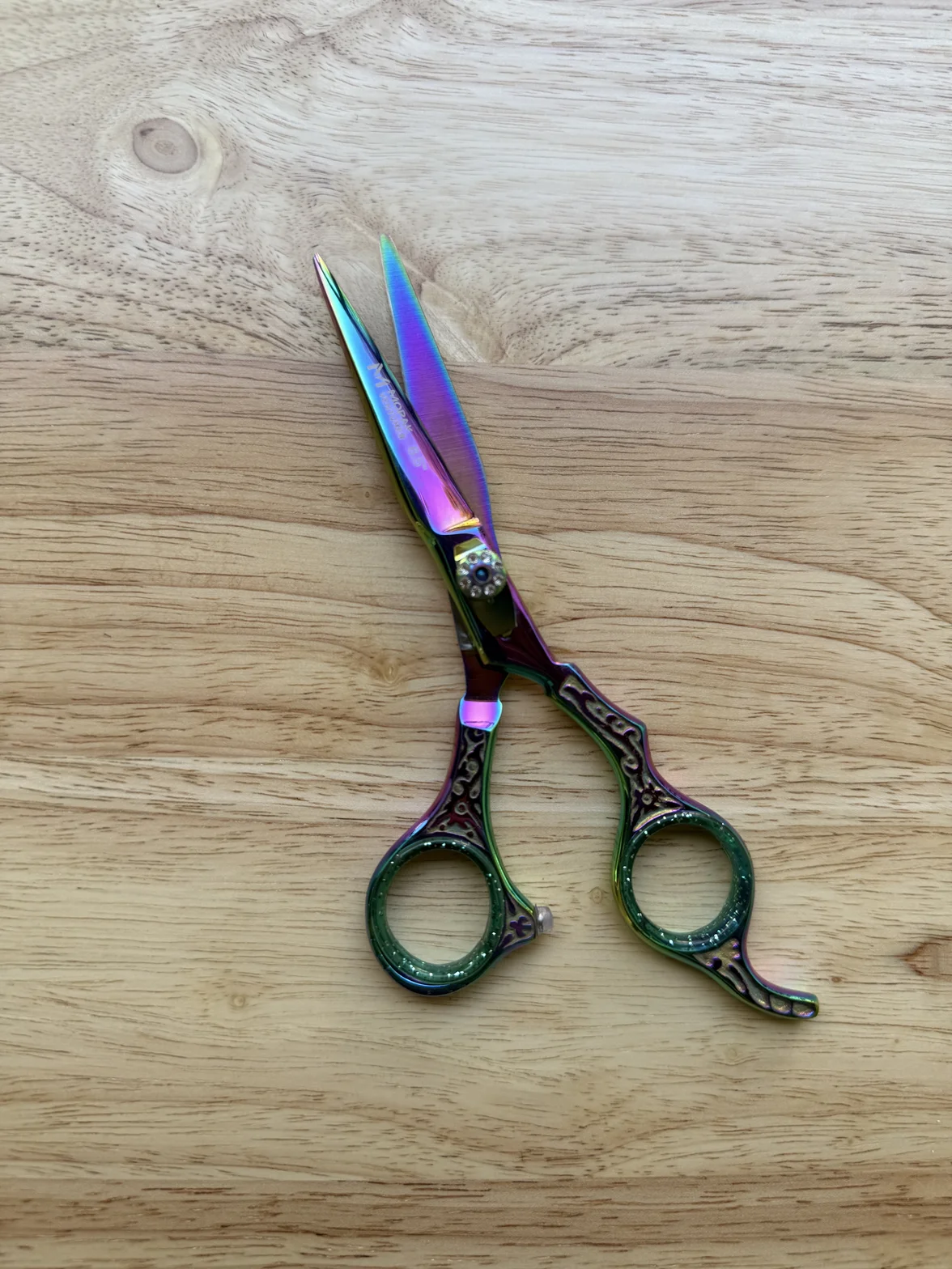 An ornate pair of iridescent professional hair-cutting scissors with decorative handles on a light wooden surface