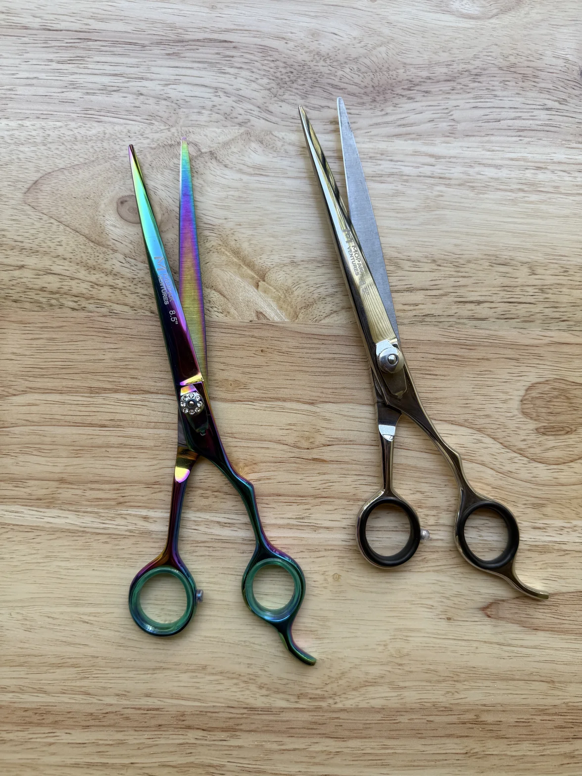 Two professional hairdressing scissors on a light wood surface, including an iridescent pair and a polished silver pair
