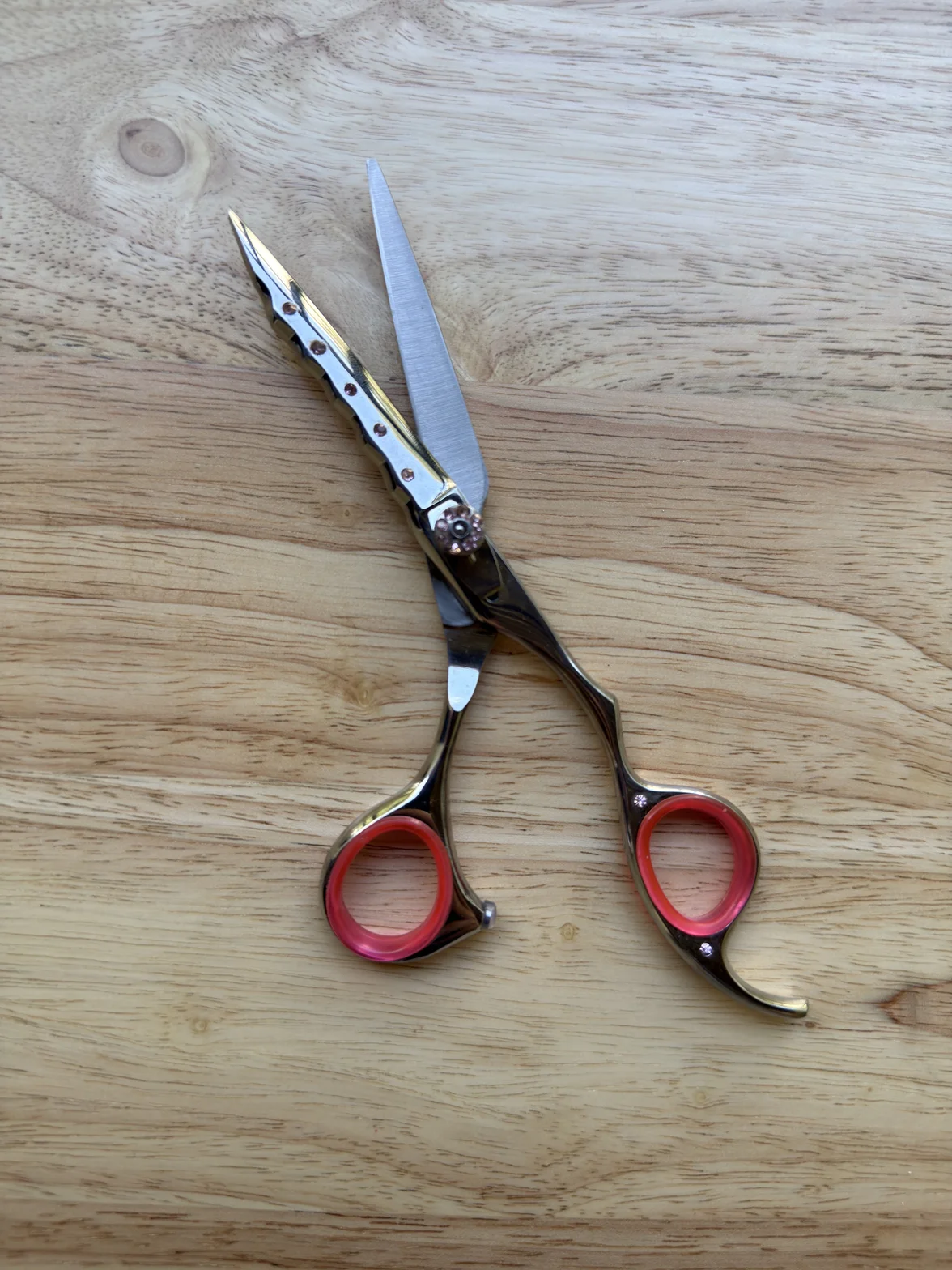 Polished professional hairdressing scissors with red finger rings, shown crossed on a light wooden surface