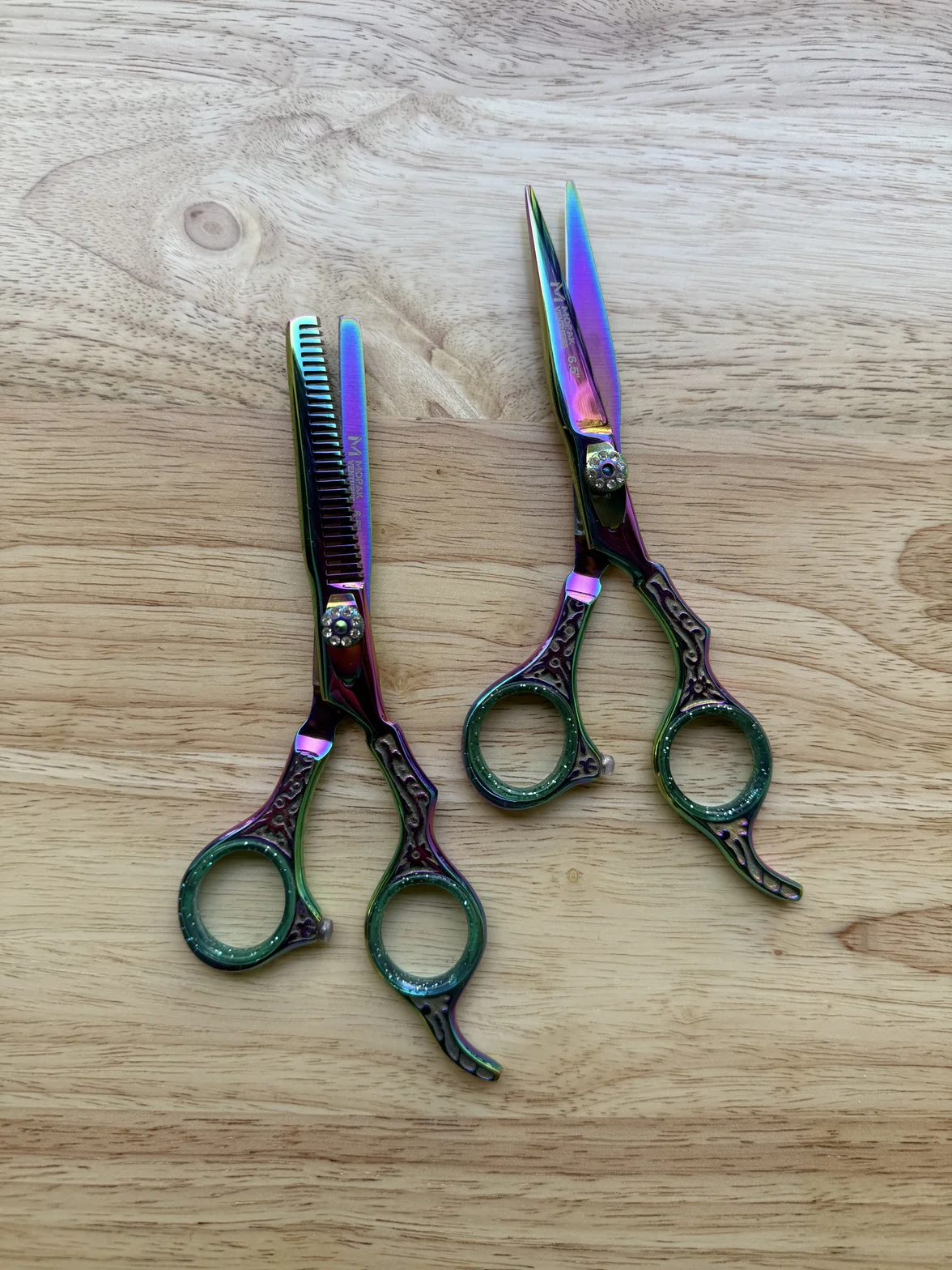 Two decorative professional hairdressing scissors, including a thinning shear, displayed on a light wooden surface