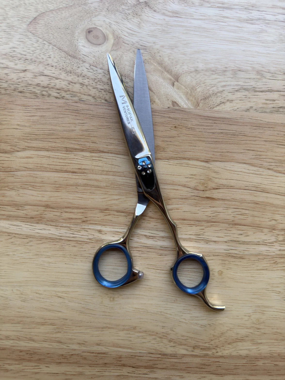 Professional hair-cutting scissors with polished metal blades, blue finger rings, and an adjustable tension screw on a light wooden surface