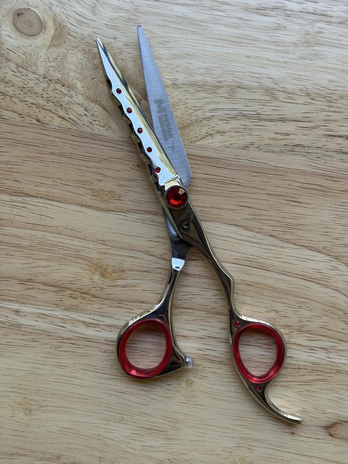 Polished professional hair-cutting scissors with red finger rings and decorative accents on a light wooden surface