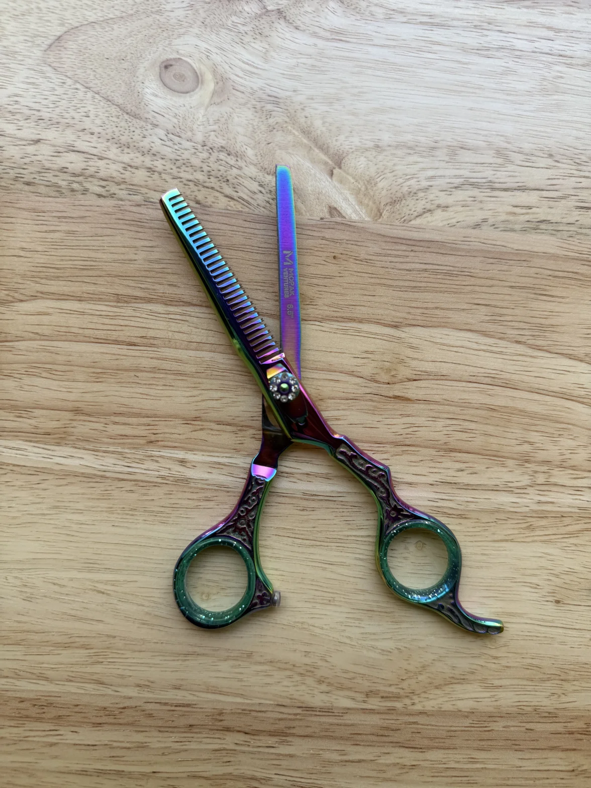 A colorful iridescent hair-cutting and texturizing shear with a comb-like blade on a light wood surface